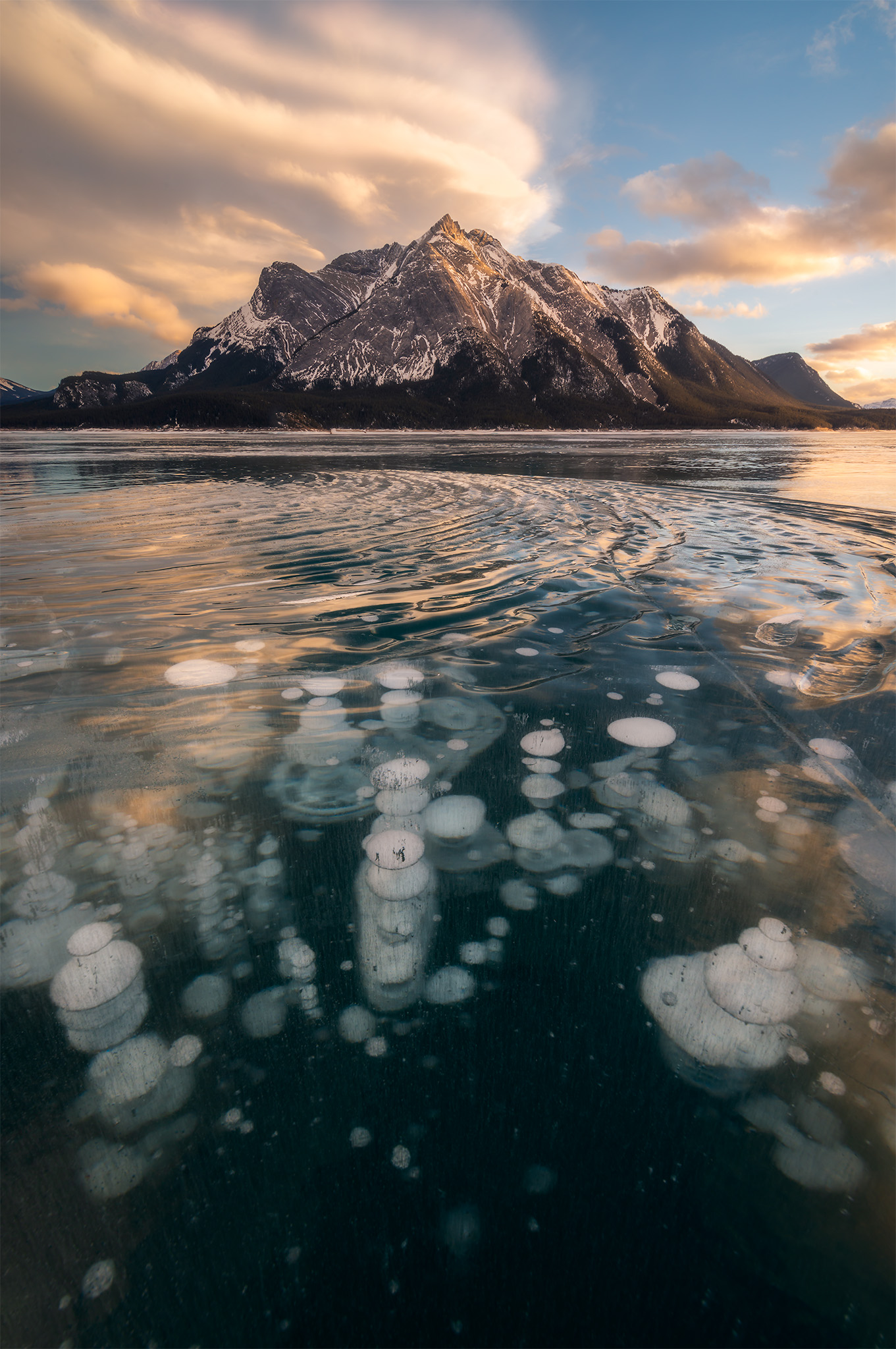 A landscape photograph of Abraham Lake at sunset with a lenticular cloud over Mount Michener and frozen methane bubbles